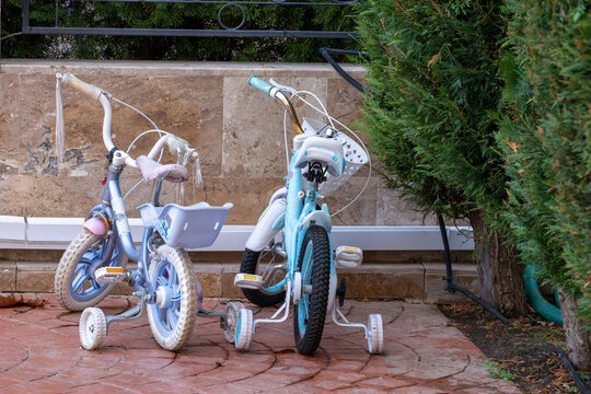 two light blue children's bicycles equipped with training wheels, parked together on a paved stone patio next to a low stone wall and a green evergreen bush.