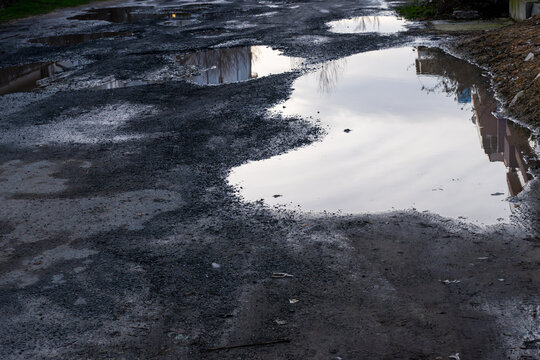 a heavily damaged and uneven dirt road riddled with large, muddy potholes filled with rainwater, reflecting the bright sky and nearby structures on the water's surface.