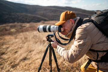 A close-up view of a wildlife photographer using tripod and aiming a large telephoto lens across an open countryside landscape on sunny day.