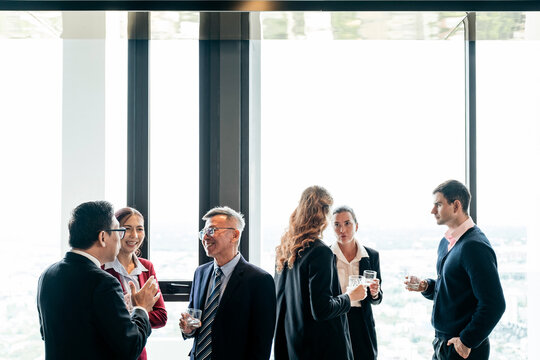 Professionals networking by large windows in a high-rise office, chatting and holding drinks with a bright city view in the background.