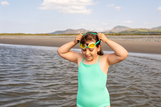 Child girl putting on swimming goggles while standing in shallow sea.