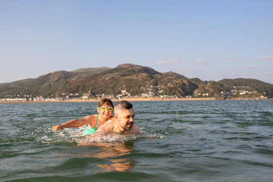 Father swimming with daughter on his back in sea.