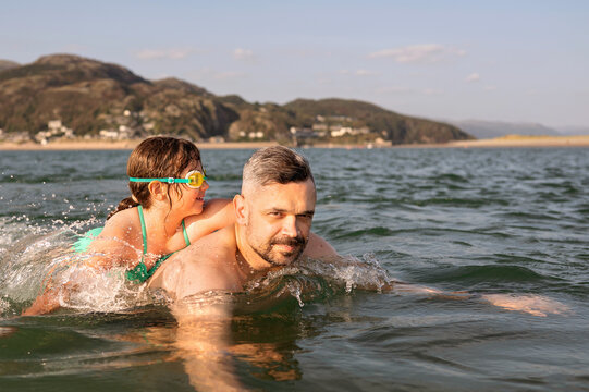 Girl holding father while swimming in coastal water.