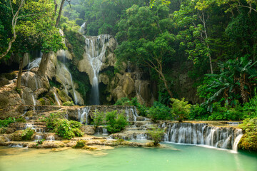 Emerald green pools collect at the base of tiered limestone cascades surrounded by dense tropical forest at Kuang Si Waterfalls in northern Laos. Sunlight filters through lush foliage, enhancing the © Florent
