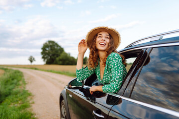 Cheerful woman leans out window, enjoying rural landscape on sunny day. Young traveler in bright...