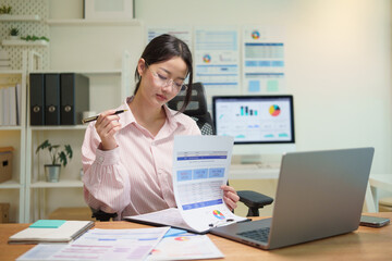 Businesswoman reviewing financial report documents while working on a laptop in a modern office.
