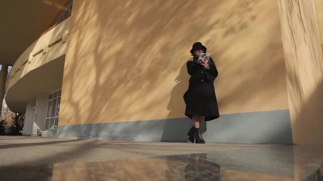 Senior woman author in black coat and hat writes notes in notebook leaning on building facade wall. Thoughtful elderly lady finishes daily plan and puts notebook into handbag outdoors