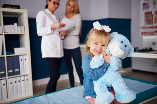 Girl hugging teddy in medical practice with doctor and mother in background