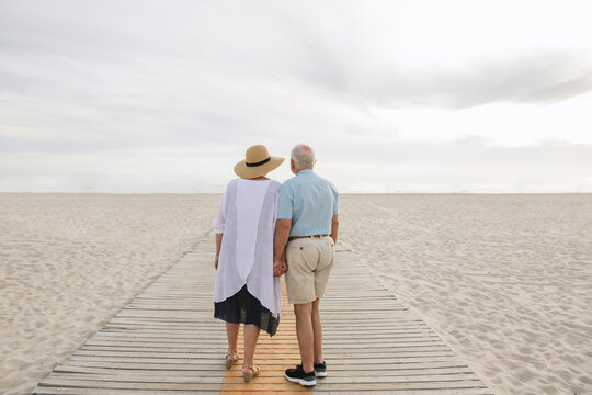 Back view of senior couple standing hand in hand on wooden boardwalk looking at horizon, Liepaja, Latvia