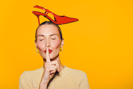Young woman with red high heel shoes on her head makes shushing gesture with finger on lips. Bright yellow background creates playful and creative mood in fashion concept.