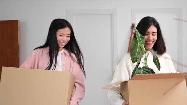 Two cheerful young women carrying cardboard boxes with belongings enter their new apartment and hug
