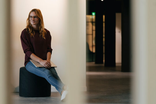 Smiling businesswoman sitting on stool holding laptop