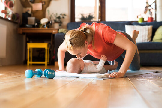 Happy mother kissing baby at home lying on mat next to dumbbells