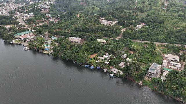 High-angle drone shot capturing the lush green landscape and residential buildings along the banks of the Volta River near the Akosombo Dam in Ghana. This footage showcases the intersection of nature