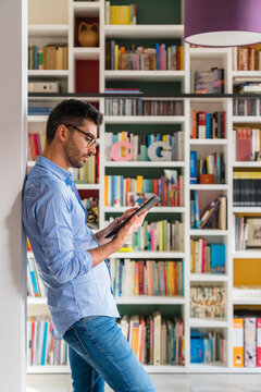Young man standing in front of bookshelves at home using digital tablet