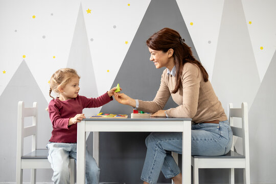Preschool teacher and young student share a geometric shape toy at a small table, engaging in playful learning and early childhood development in a bright, cheerful classroom setting