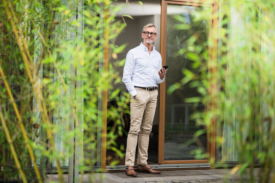 Senior man with grey hair standing in front of his modern design home holding smartphone