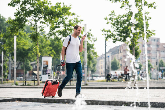 Man with rolling suitcase and cell phone walking at fountain in the city