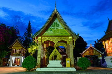 Obraz premium An intricately decorated Buddhist pavilion glows under artificial lights against a vivid blue evening sky in Luang Prabang, Laos. Golden patterns and traditional architectural details stand out