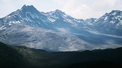 Towering mountains with snow covered peaks dominate the horizon, casting deep shadows on the rocky terrain. The sky is overcast yet bright, offering a serene and majestic landscape.