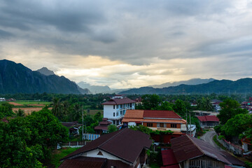Fototapeta premium A cluster of red-roofed houses and a white building sit among lush greenery in Vang Vieng, Laos, with dramatic limestone mountains and layered hills in the distance. Moody evening clouds and soft