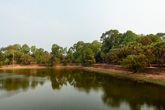 A tranquil moat edged with stacked stone embankments and lush palm trees near Bakong in Cambodia. The calm water reflects the greenery and earthy banks, creating a serene tropical landscape under soft