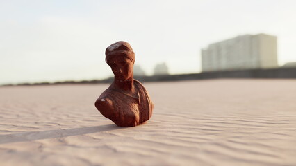 A small, weathered sculpture stands on a sandy surface as the sun sets behind a city skyline. The warm glow highlights the intricate details of the figure, suggesting a rich history. © icetray