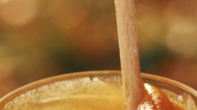 Close-up detailed view of a beekeeper's hand stirring creamy white rapeseed honey in a glass jar at the apiary, showcasing the smooth texture