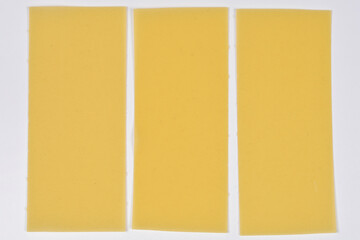 Three rectangular sheets of dry yellow lasagna pasta arranged side by side on a plain white...