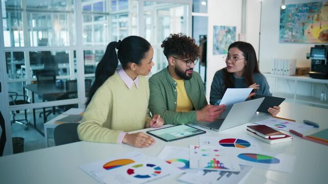 Wide view of corporate team planning campaign. Three colleagues studying printed charts. Male comparing laptop data. Woman holding tablet and smiling. Brainstorming marketing strategy in glass office.
