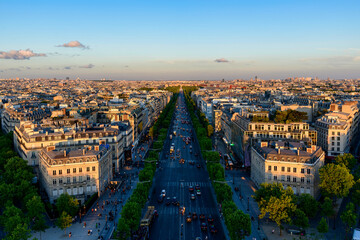 Obraz premium Wide aerial view of the Avenue des Champs Elysees stretching through central Paris, lined with elegant Haussmann buildings and green trees in warm evening sunlight under a clear blue sky.