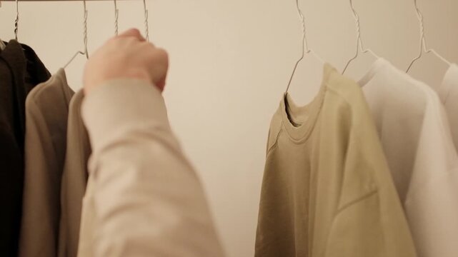 Young woman selects or organizes clothes on a clothing rack. Choosing pastel color knitwear t-shirts and sweatshirts. New basic casual unisex clothes. Concept of fast fashion industry waste issue. 