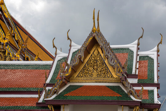 Close-up of the multi-tiered roofs of a temple building at the Grand Palace. The roofs are covered in green and orange glazed tiles and feature ornate golden pediments with traditional carvings.