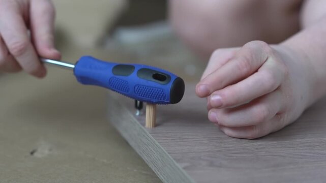 A teenager carefully inserts wooden dowels into pre-drilled holes on a white furniture panel and firmly hammers them into place with a screwdriver handle