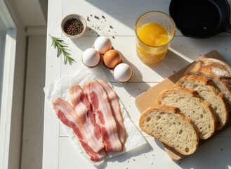 Overhead view of a delicious breakfast spread with crispy bacon farm fresh eggs sliced artisan bread and a glass of orange juice on a rustic white table with natural sunlight casting shadows