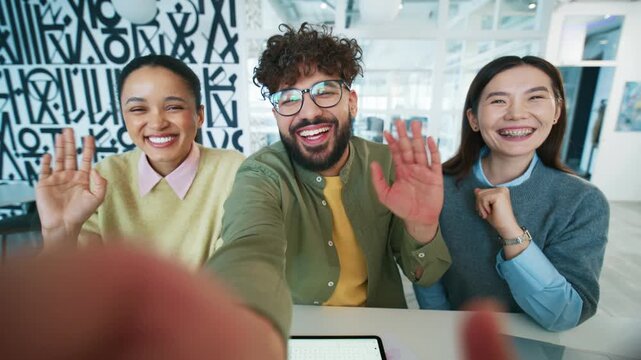 Happy team posing for group selfie at desk. Colleagues holding camera and extending arm. Business people waving hands and laughing. Friendly workers greeting viewer inside modern office.