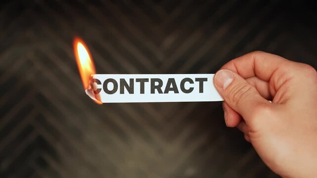 Close-up of a hand holding a paper strip labeled &ldquo;CONTRACT&rdquo; while the edge burns, symbolizing agreement risk, breach, termination, and legal fallout on a dark background with copy space.