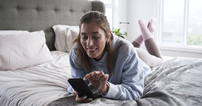 Glancing at phone screen, middle-aged woman is scrolling on bed in pink socks chuckling