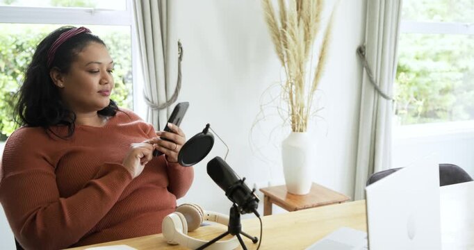 Mid adult woman scrolling phone, glancing at microphone and preparing to record at desk by window