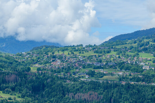 Verdant green hills dotted with charming alpine houses and dense forests surround the village of Combloux, set beneath dramatic cumulus clouds and a vibrant blue sky.