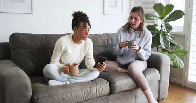 Zooming in, Diverse female friends wearing eye patches scrolling phone and sipping coffee at home