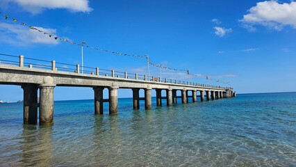 Concrete pier with colorful flags extending over crystal clear turquoise waters in Porto Santo, Madeira, Portugal, beneath a vivid blue sky.