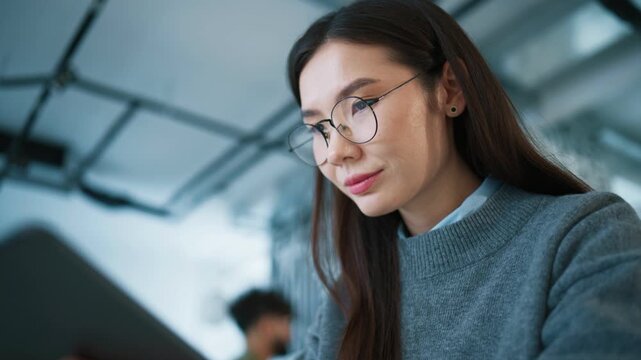 Modern classroom interior. Asian female student wearing glasses holding tablet with one hand. Reading lecture slides on screen. Focused expression. Studying course materials and preparing for exam.