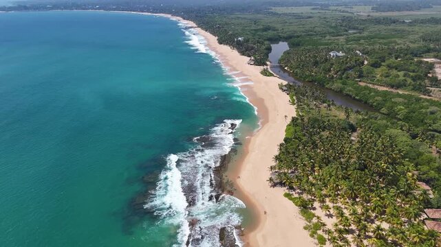 Breathtaking aerial drone footage of the vast coastline in Tangalle, Sri Lanka. The video captures a long stretch of golden sandy beach separating the turquoise Indian Ocean waves from a calm river