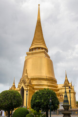 Fototapeta premium Closeup of the gold leafed stupa (Phra Siratana Chedi) within the Grand Palace complex in Bangkok, Thailand. The spire reaches toward a cloudy, overcast sky, reflecting a warm glow.