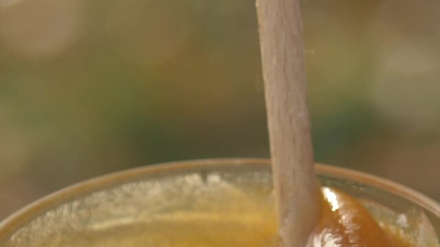 Close-up detailed view of a beekeeper's hand stirring creamy white rapeseed honey in a glass jar at the apiary, showcasing the smooth texture