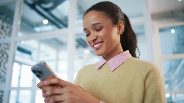 Professional woman holding smartphone and smiling at screen. Standing inside corporate building near glass partitions. Female chatting online with someone close. Digital communication concept.