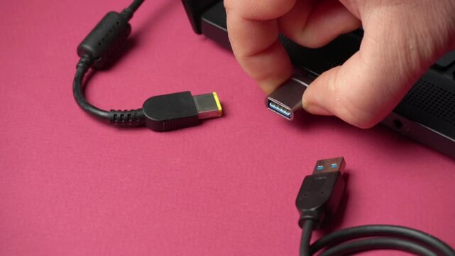 Hand connecting USB cable to laptop port, showcasing the progression of plugging in a device with visible cables and connectors on a pink background