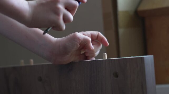 A teenager carefully inserts wooden dowels into pre-drilled holes on a white furniture panel and firmly hammers them into place with a screwdriver handle
