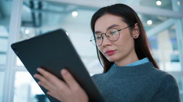 Female employee holding tablet and entering text with one hand while standing in glass office corridor. Woman wearing glasses concentrating on digital project inside corporate workspace.